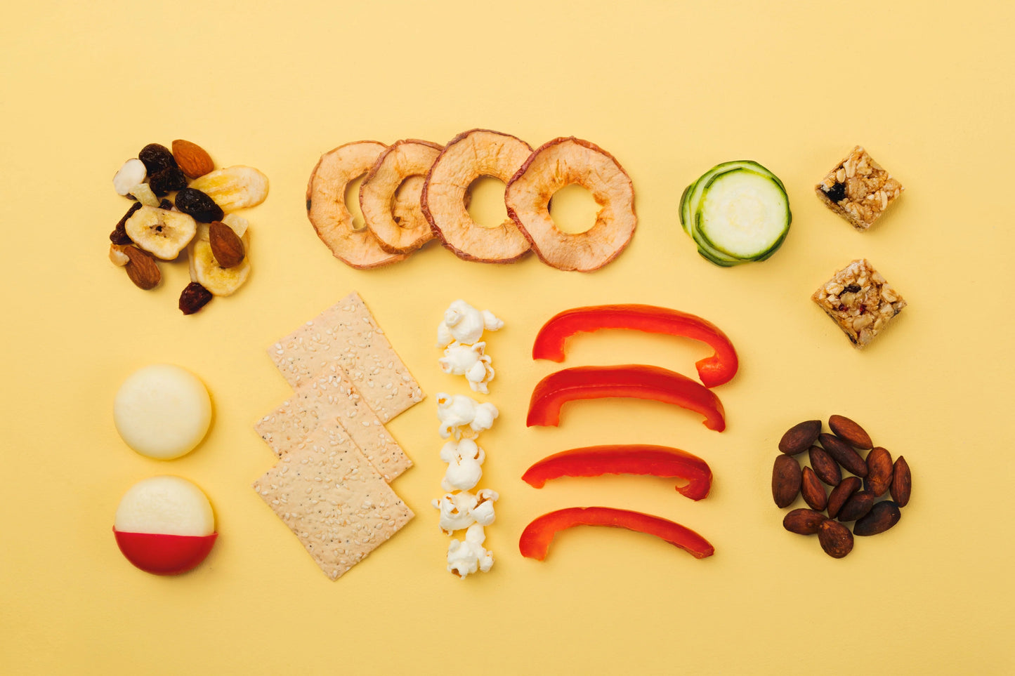 Gelber Hintergrund, davor verschiedene Lebensmittel für das Pausenbrot (Paprika, Apfelringe, Nüsse, Gurke)